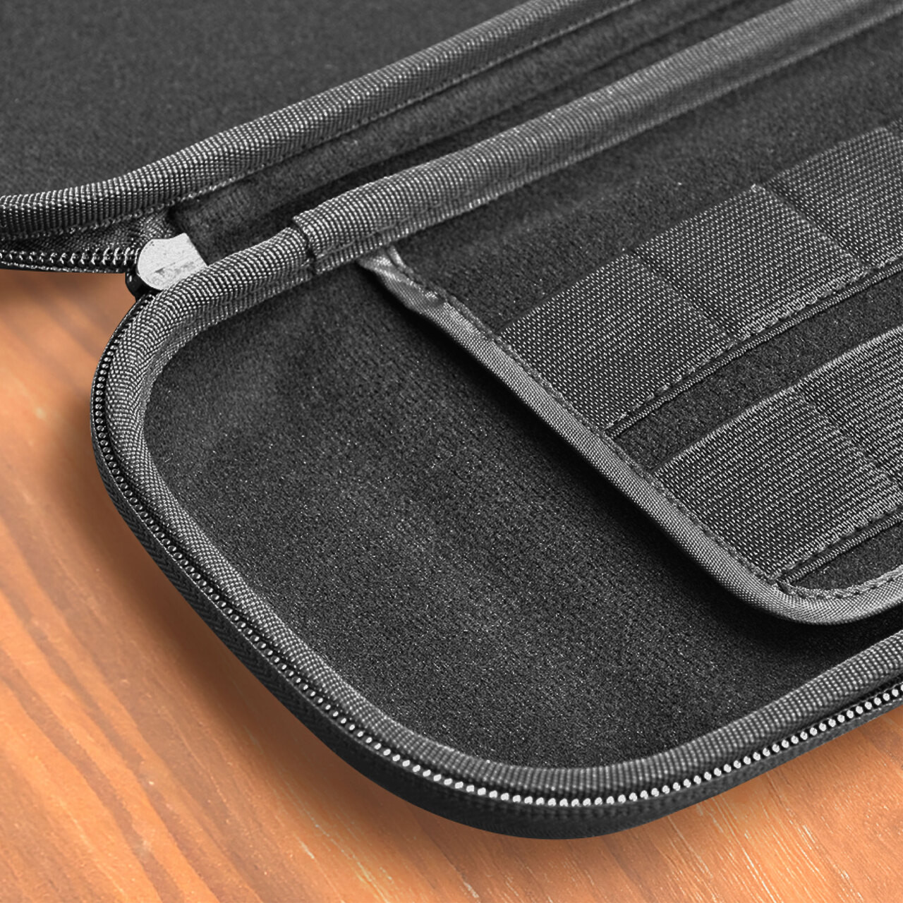 Primo piano di una custodia nera con cerniera, aperta per rivelare scomparti interni. La custodia poggia su una superficie di legno. I dettagli includono cuciture e tessuto.