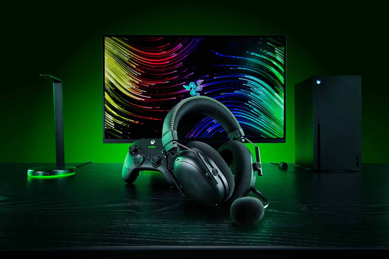 Gaming-Setup. Razer-Headset, Xbox-Controller, Konsole, Monitor mit buntem Hintergrund, alles auf einem dunklen Schreibtisch.