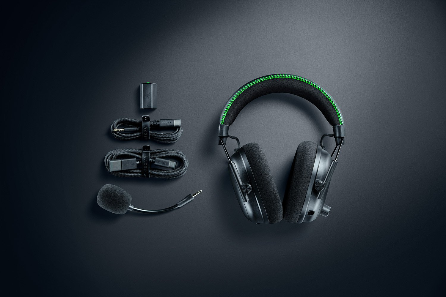 Schwarzes Gaming-Headset mit grünen Akzenten, Kabeln, Mikrofon und einer Ladekomponente auf dunkler Oberfläche.
