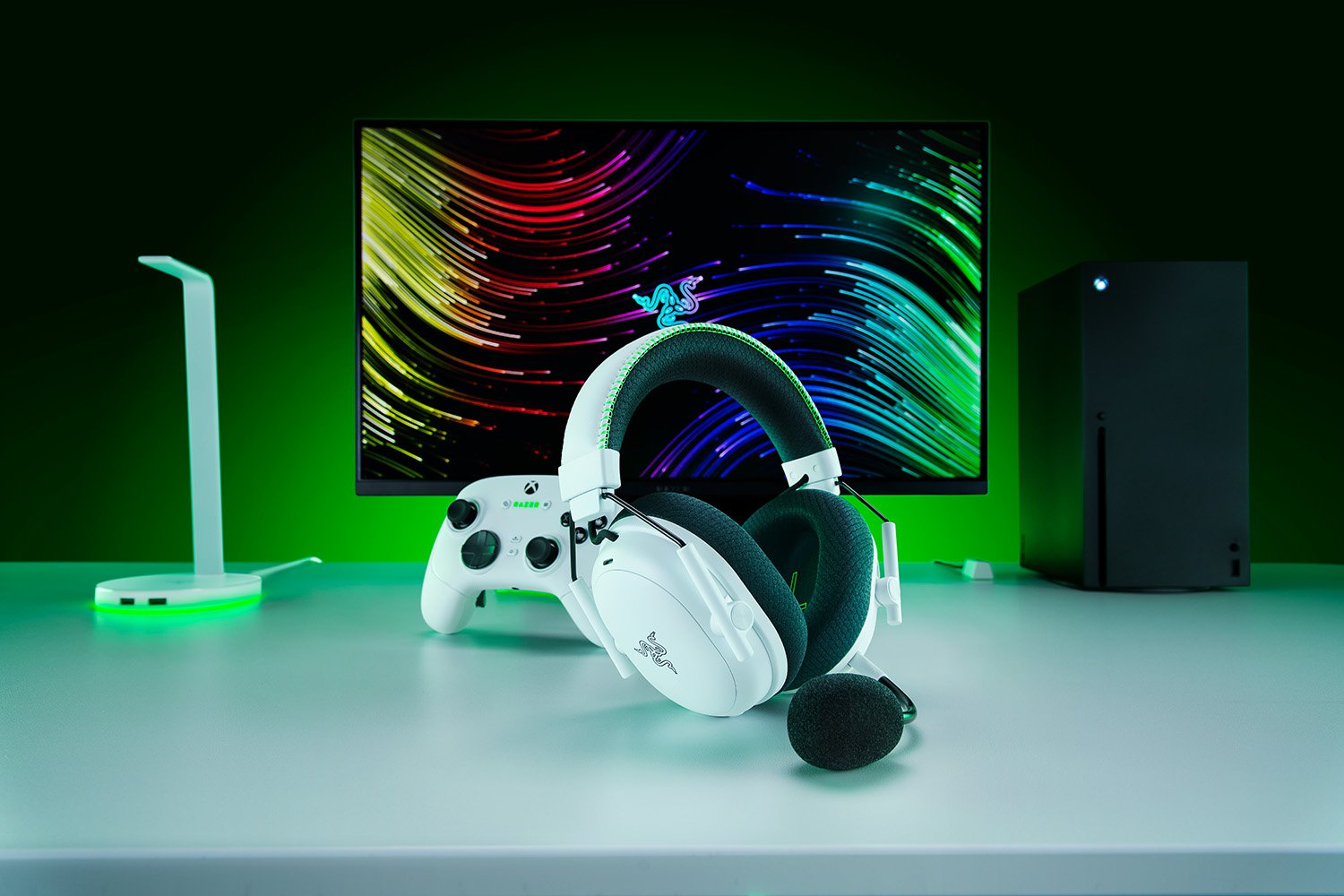 Casque de jeu Razer blanc, manette Xbox et moniteur avec fond coloré sur un bureau blanc.