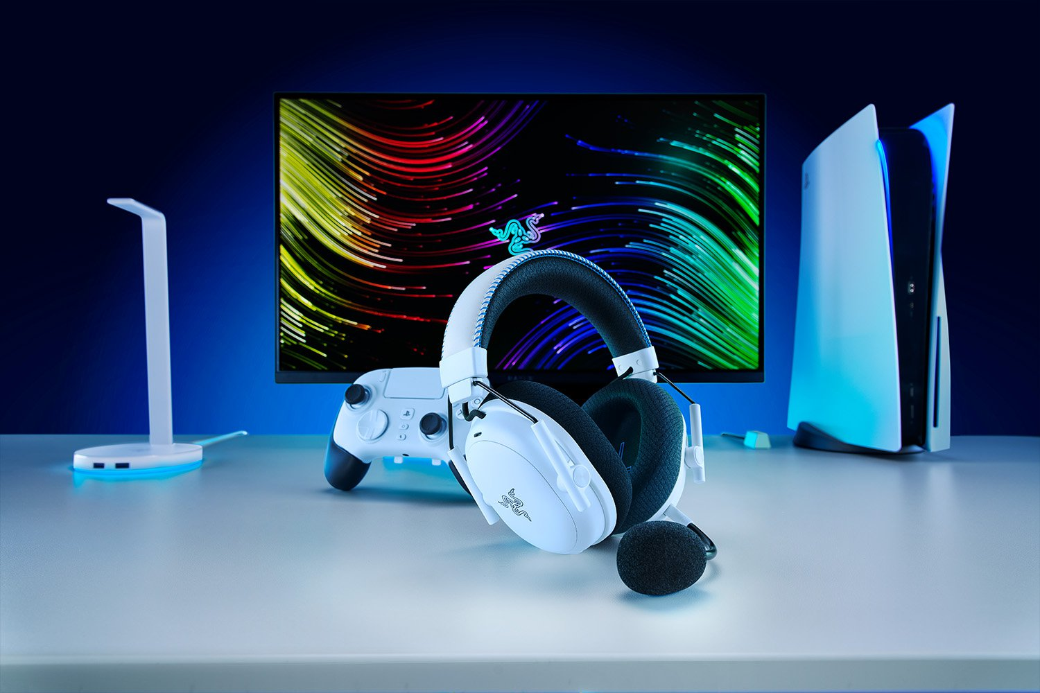 Weiße Gaming-Kopfhörer, Controller und PS5-Konsole auf einem Schreibtisch. Ein Monitor zeigt farbige Linien, links eine Schreibtischlampe.