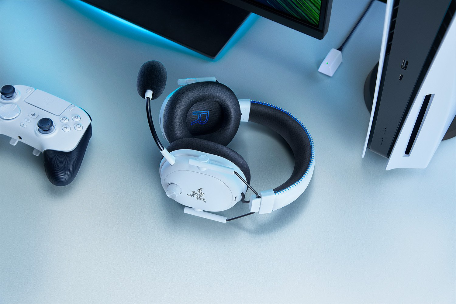 Weißes Gaming-Headset, Controller und PS5-Konsole auf einem Schreibtisch. Blaue Akzente, Razer-Logo sichtbar.