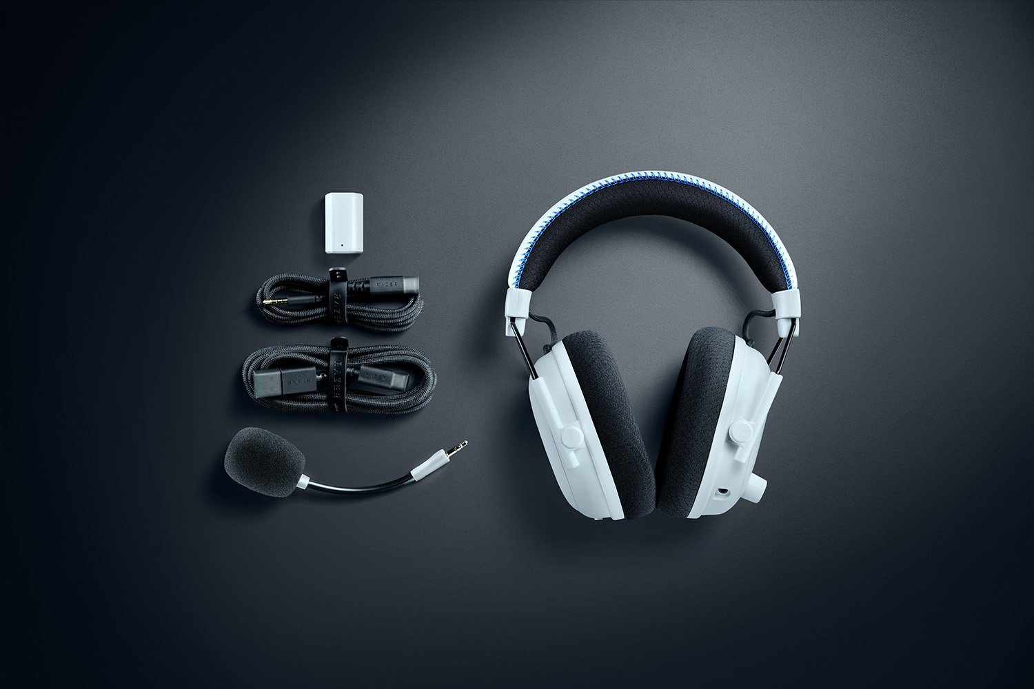 Weißes Headset mit schwarzen Ohrpolstern, Mikrofon, Kabeln und einem kleinen weißen Gerät auf dunkler grauer Oberfläche. Zubehör sichtbar.