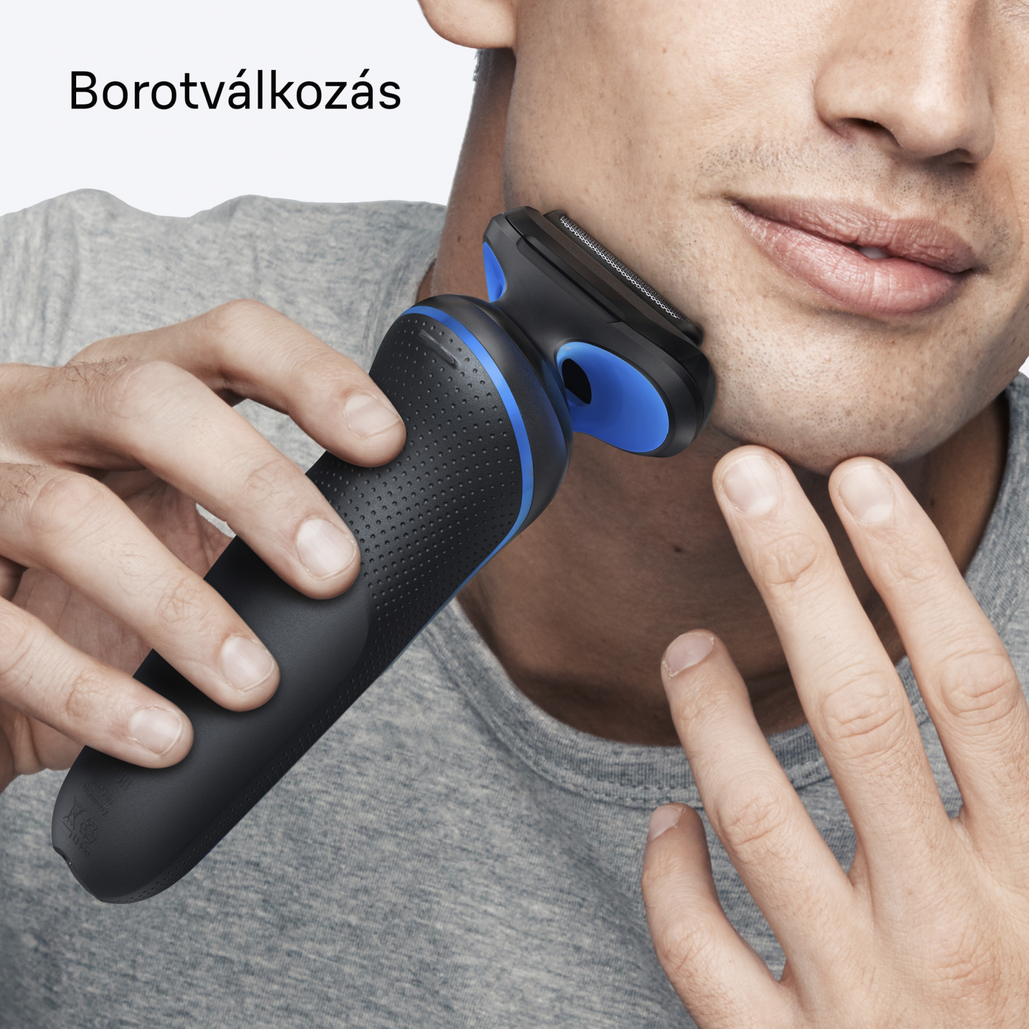 Egy férfi elektromos borotvával borotválkozik. A borotva fekete és kék. A férfi szürke inget visel.