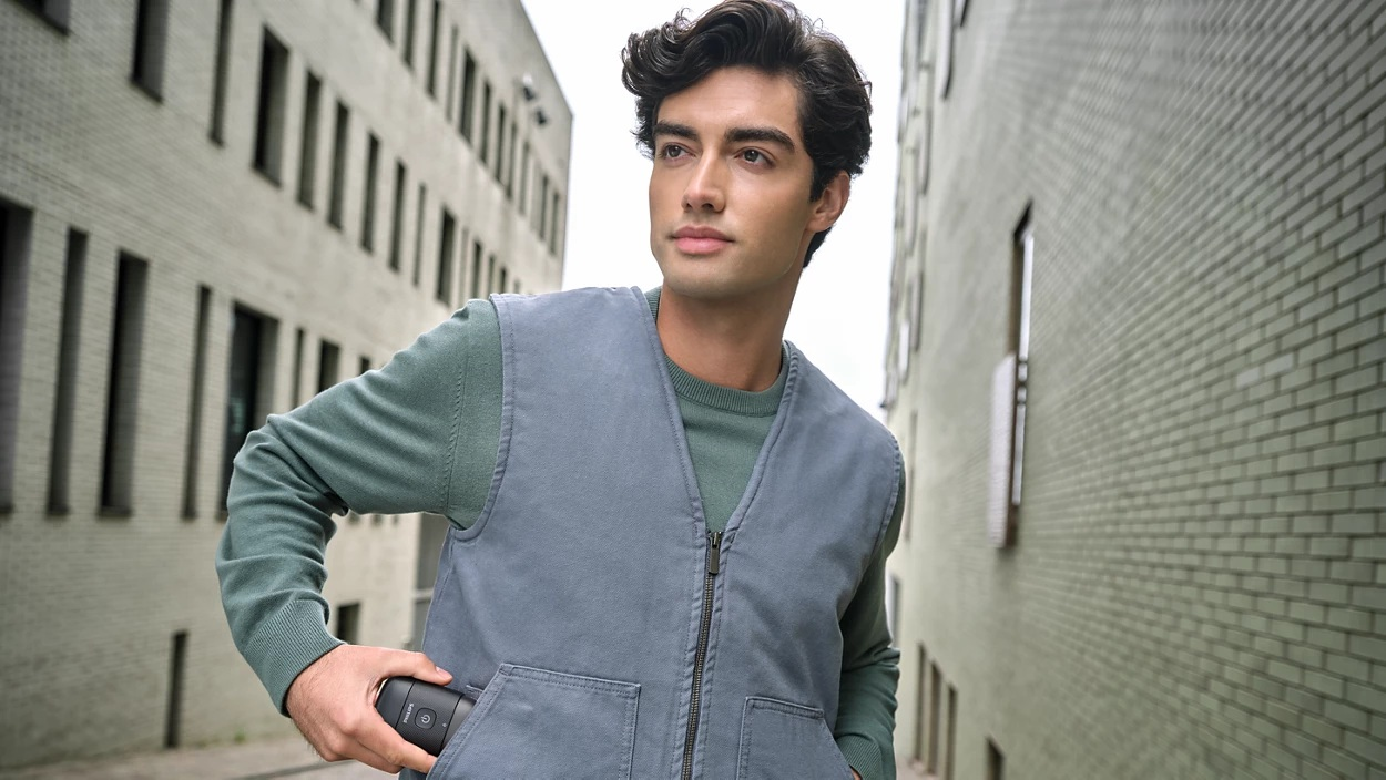 Mężczyzna w niebieskiej kamizelce trzyma golarkę. Budynki w tle. Patrzy w górę i na bok.