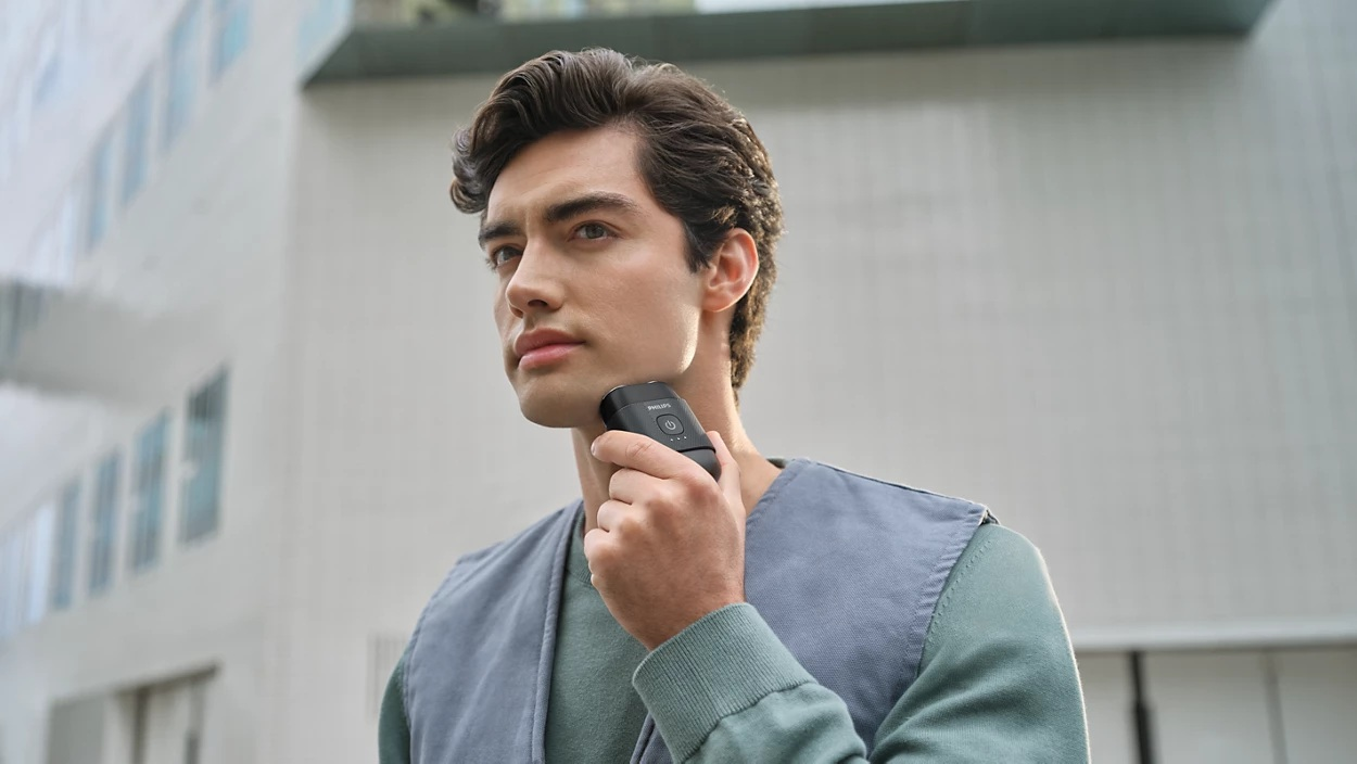 Mężczyzna goli twarz czarną golarką elektryczną. Ma na sobie niebieską kamizelkę i zieloną koszulę. Tło to budynek.