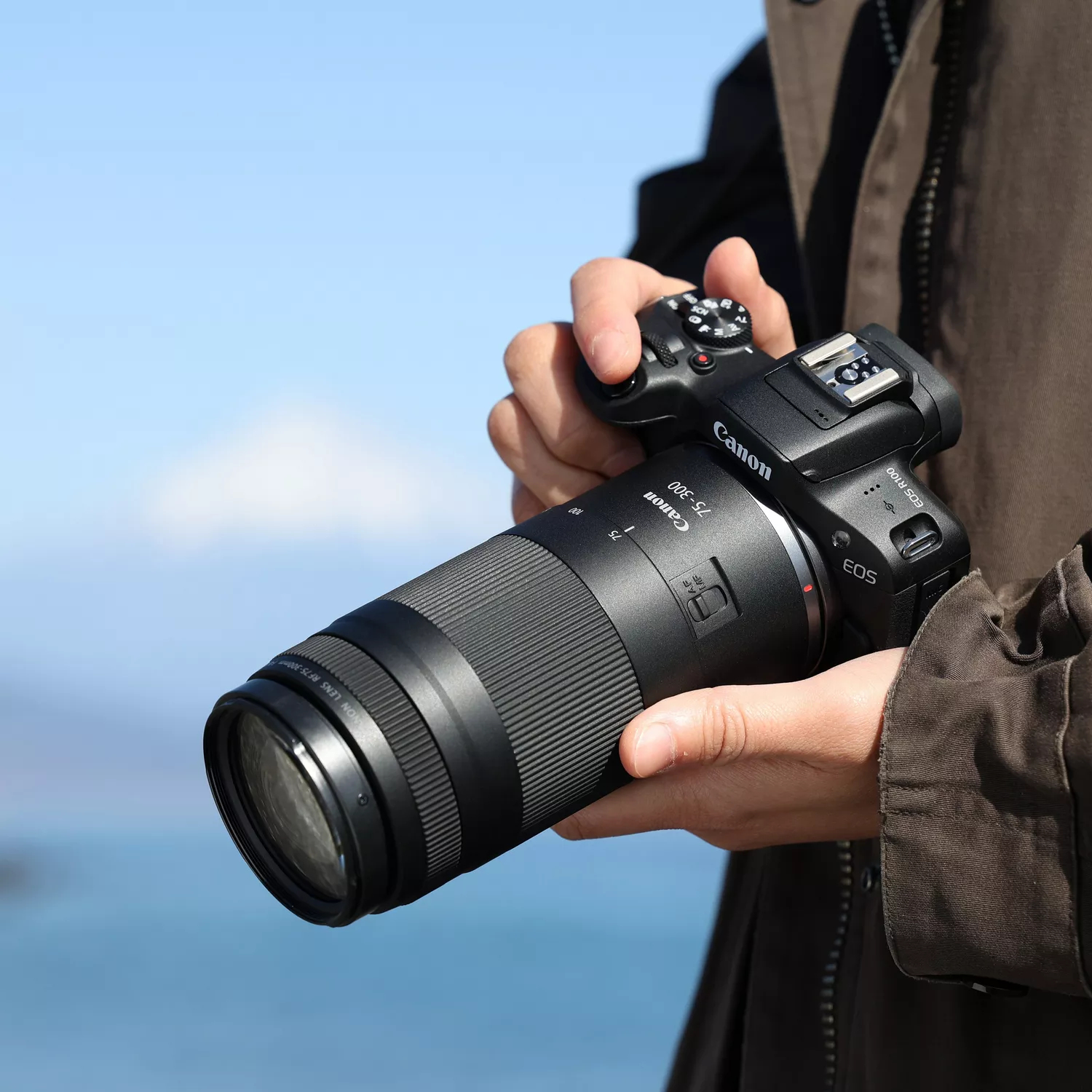 Eine Person hält eine Canon-Kamera mit einem langen Objektiv vor blauem Himmel. Die Kamera ist schwarz.