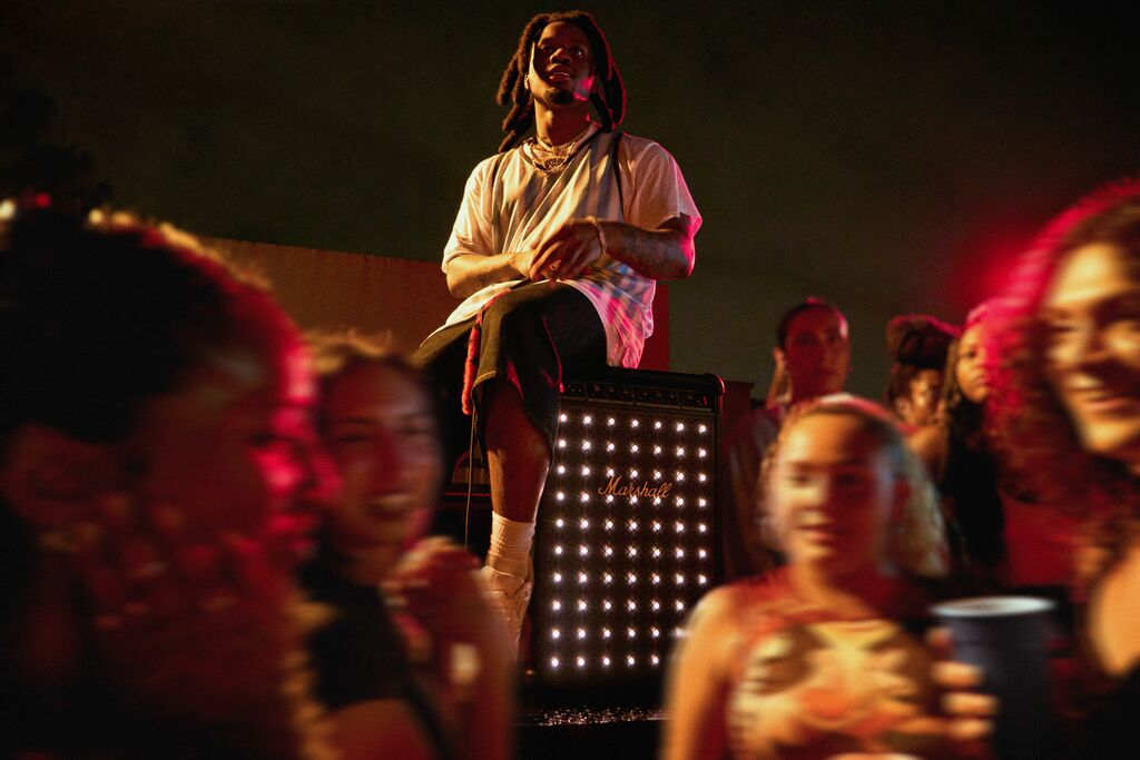Dreadlock'lu bir adam amfinin üzerinde oturuyor, etrafında insanlar var. Kırmızı aydınlatma konser atmosferi yaratıyor.