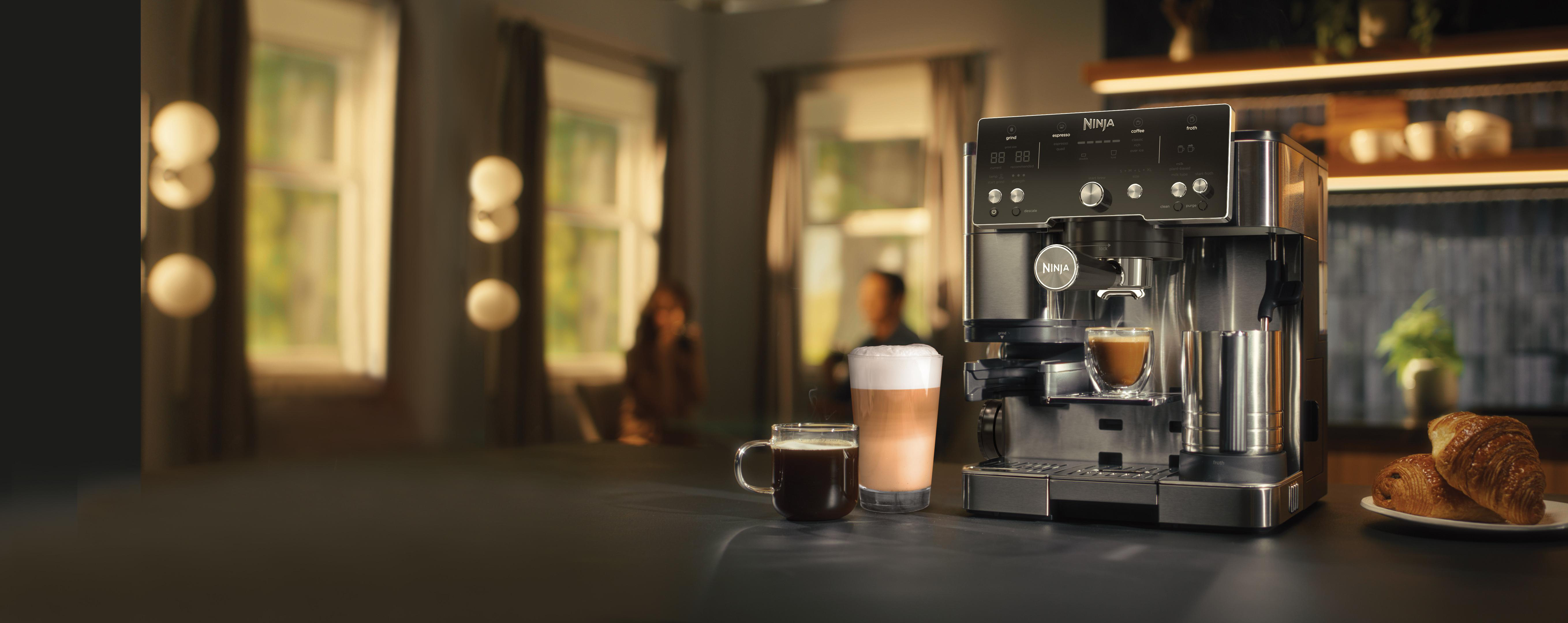Eine Ninja-Kaffeemaschine steht auf einer Theke mit Kaffeegetränken und Croissants. Fenster sind im Hintergrund.
