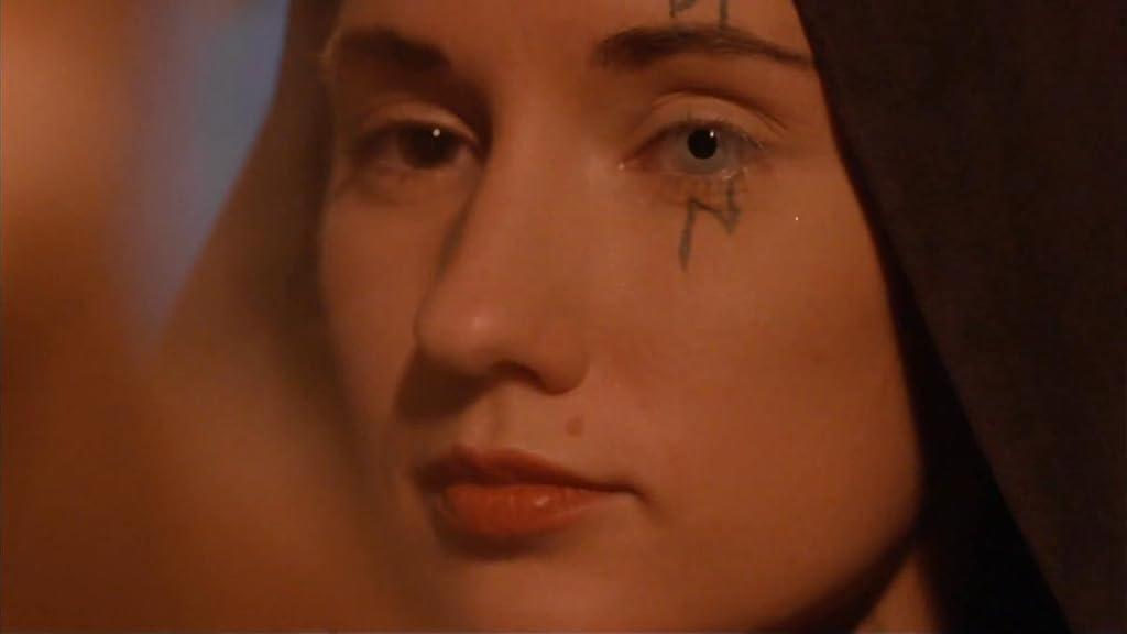 Das Gesicht einer Frau mit dunkler Kapuze. Ein Auge ist normal, das andere blass mit Tattoo darunter. Rote Lippen und ein Muttermal.