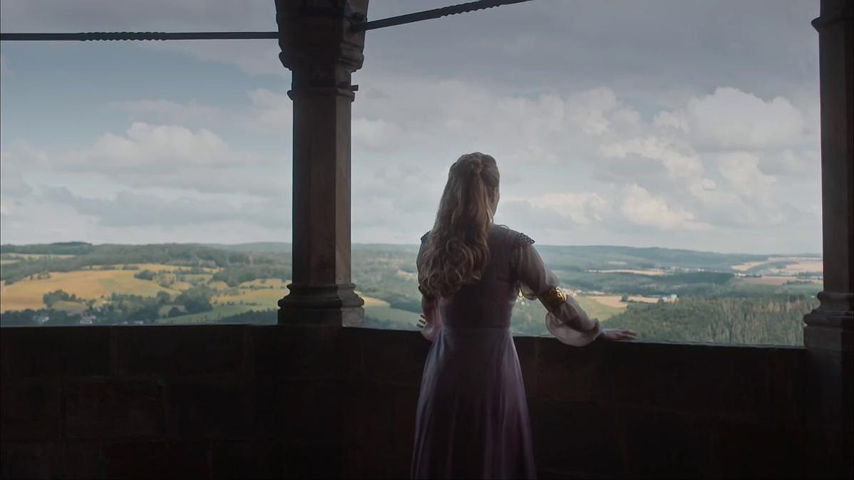Eine Frau im Kleid blickt von einem Balkon auf eine Landschaft. Hügel, Bäume und Himmel mit Wolken sind sichtbar.