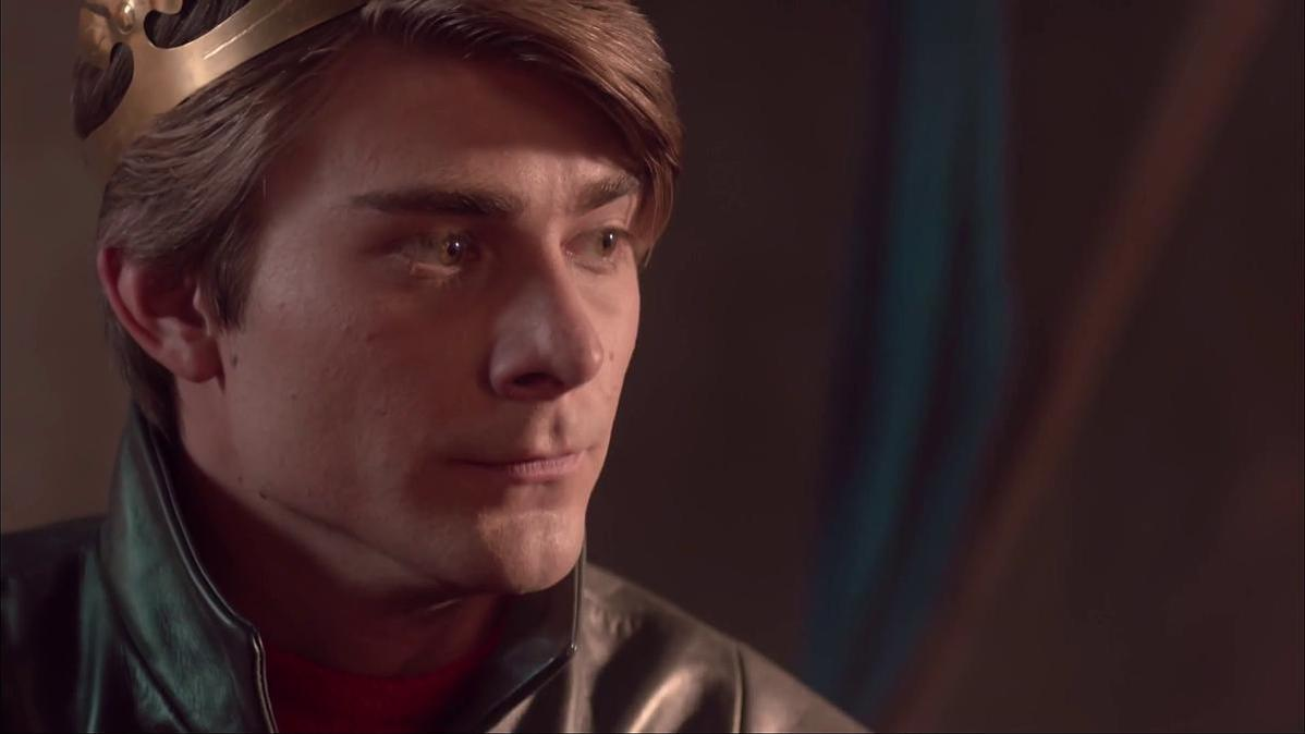 Ein Mann mit Krone schaut zur Seite. Er trägt eine schwarze Lederjacke und ein rotes Hemd. Der Hintergrund ist verschwommen.