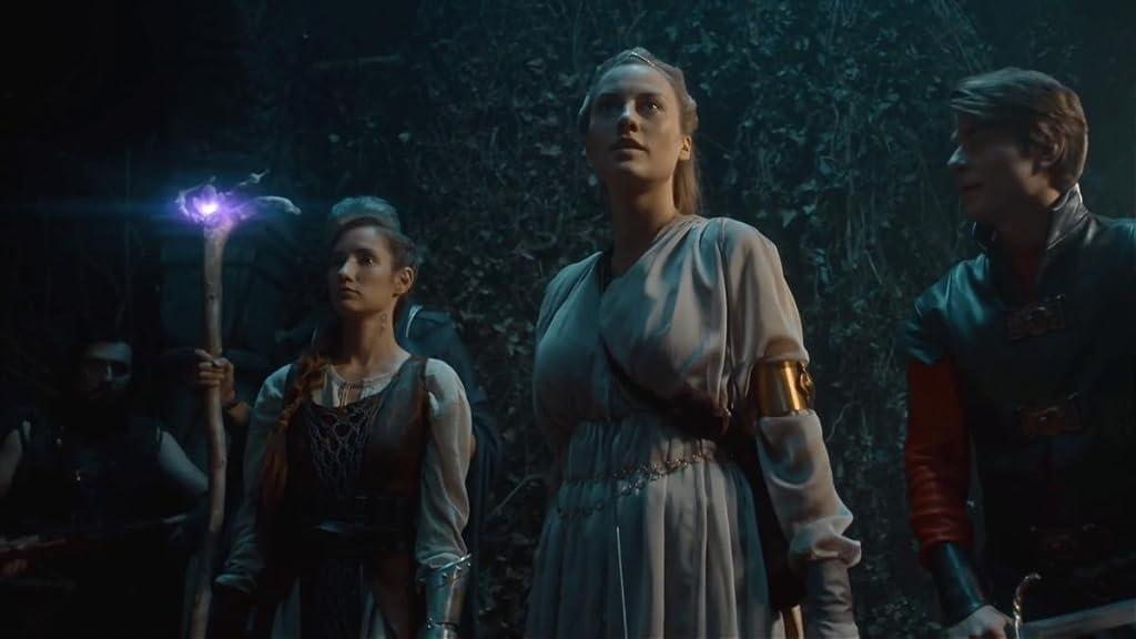 Gruppe von Menschen in Fantasy-Kleidung. Eine Frau hält einen Stab mit einem leuchtenden, violetten Orb. Waldboden. Dunkle Szene.