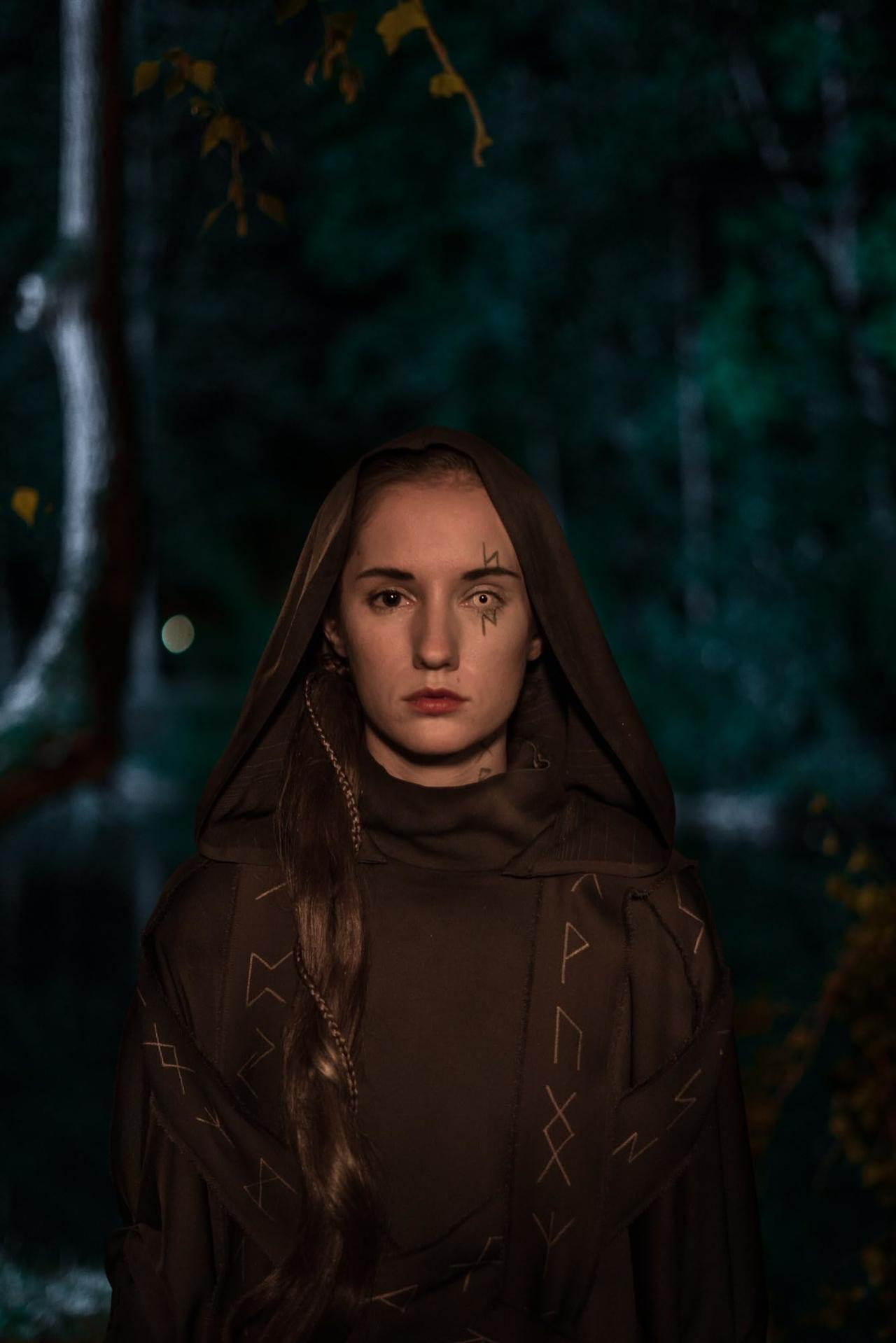 Eine Frau in einer Kapuzenrobe steht in einem Wald. Runen sind auf ihrem Gesicht und ihrer Robe. Der Hintergrund ist ein verschwommener Wald.