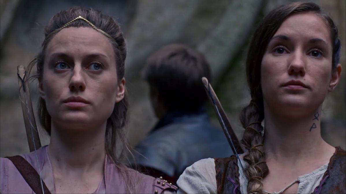 Zwei Frauen mit Pfeil und Bogen. Eine trägt ein goldenes Diadem, die andere hat ein Tattoo am Hals. Ein Mann ist verschwommen im Hintergrund.