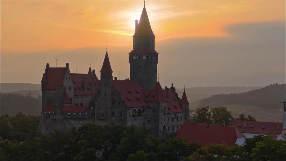Eine Burg mit roten Dächern und mehreren Türmen vor einem Sonnenuntergang. Bäume im Vordergrund, Hügel im Hintergrund.