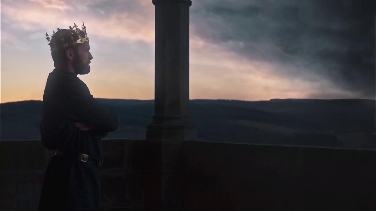 Ein Mann mit Krone steht auf einem Balkon und blickt auf einen dramatischen Sonnenuntergang über einer Landschaft.