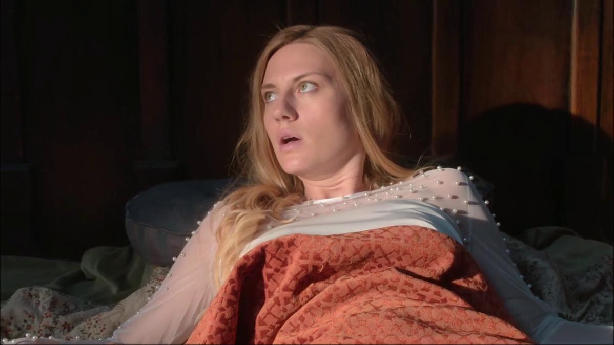 Eine blonde Frau liegt im Bett und blickt überrascht nach oben. Sie trägt ein weißes Oberteil und eine orangefarbene Decke.