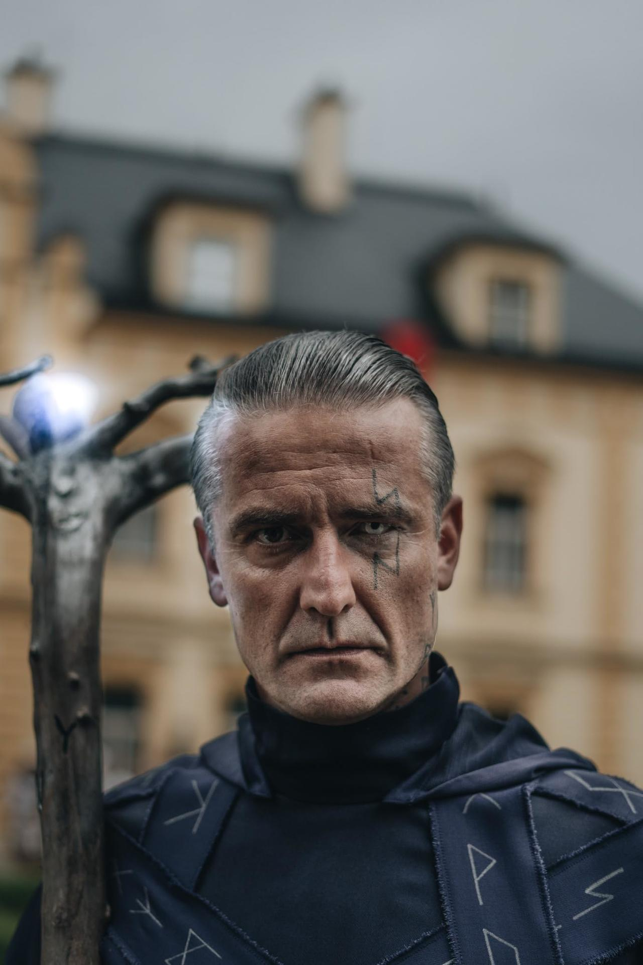 Ein Mann mit Narbe und Stab steht vor einem Gebäude. Er trägt einen dunklen Umhang. Der Stab hat eine leuchtende Kugel.