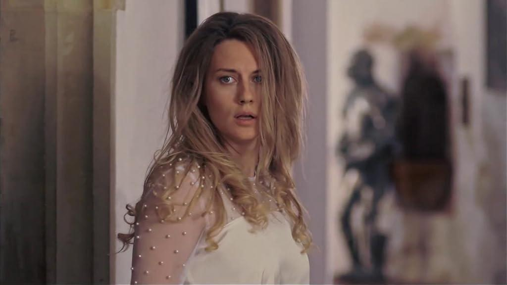 Eine Frau mit blonden Haaren und einem weißen Kleid sieht überrascht aus. Im Hintergrund steht eine verschwommene Figur.