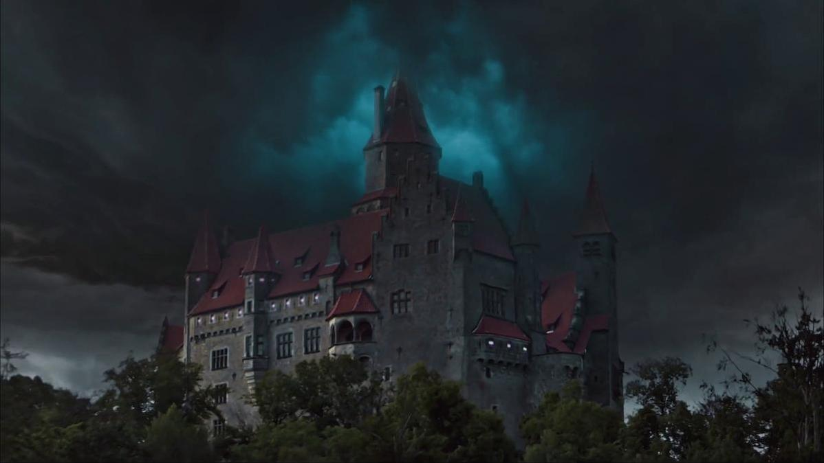 Ein großes Schloss mit roten Dächern und mehreren Türmen vor einem dunklen, stürmischen Himmel. Bäume umrahmen das Schloss im Vordergrund.
