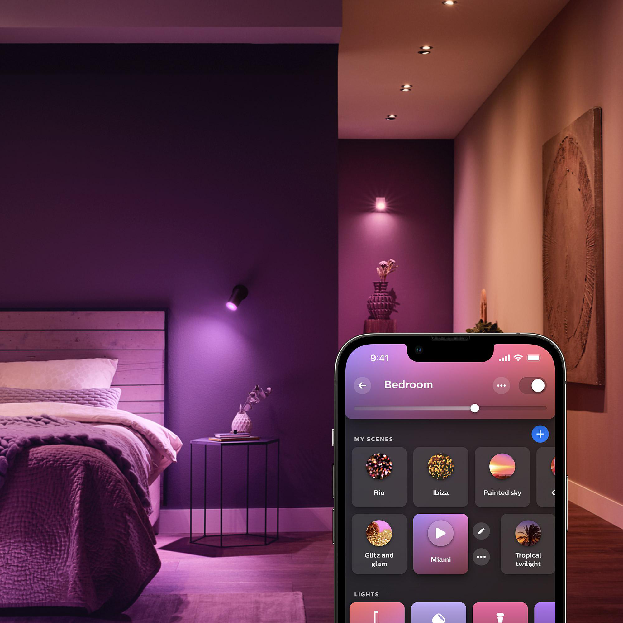 Ein Schlafzimmer mit intelligenter Beleuchtung, die über ein Telefon gesteuert wird. Bett, Beistelltisch und Wandkunst sichtbar. Lila und rosa Farbtöne.