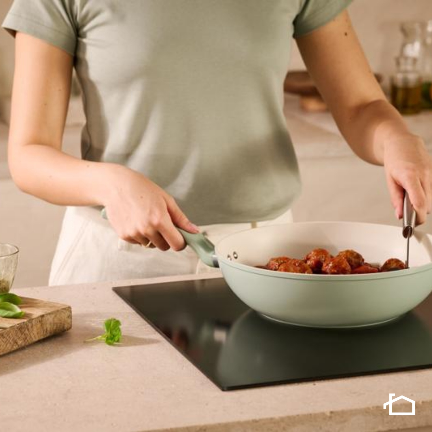 Una persona sta cucinando polpette in una padella su un fornello. L'ambiente della cucina include un tagliere e foglie di basilico.