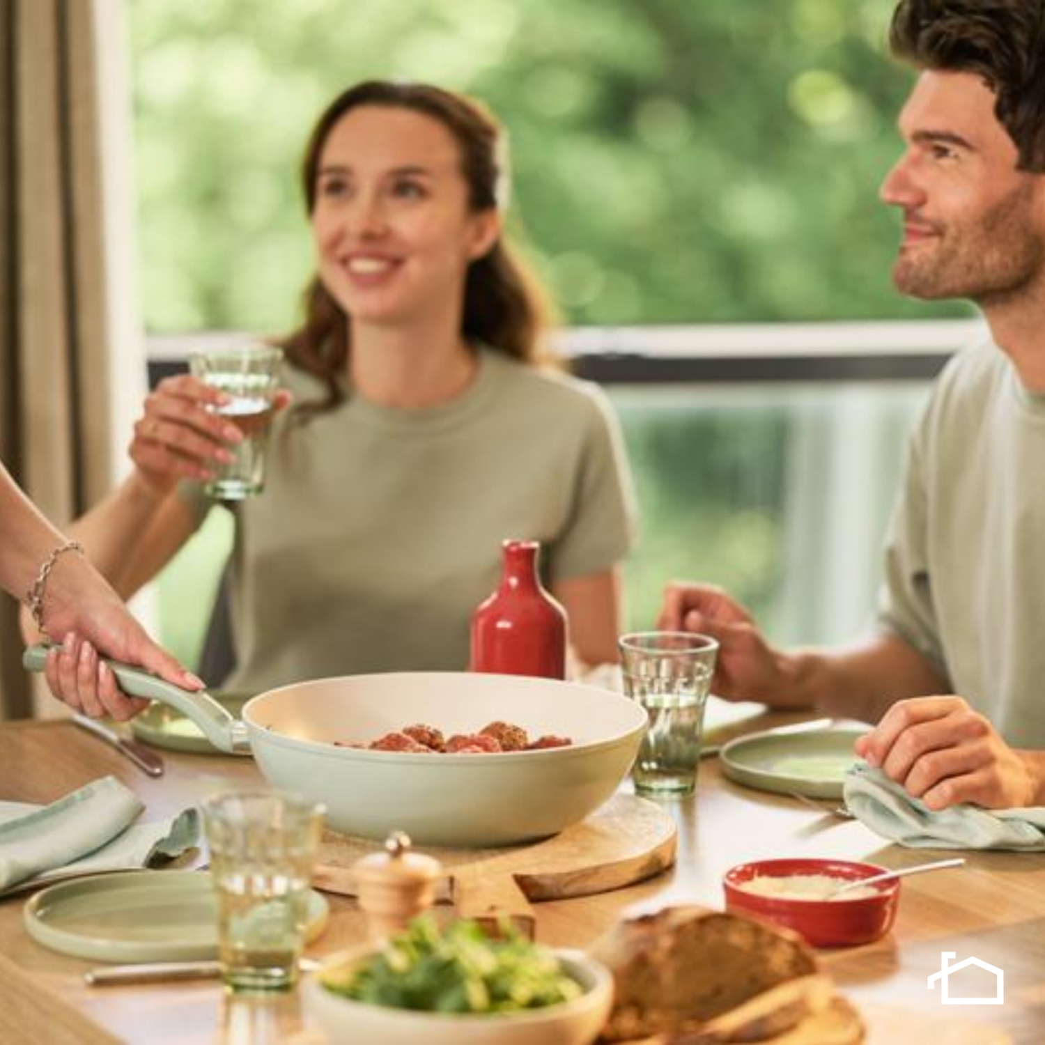 Persone a tavola. Una donna tiene un bicchiere. Il cibo viene servito in ciotole e in padella. Una bottiglia rossa e una tavola di legno sono sul tavolo.