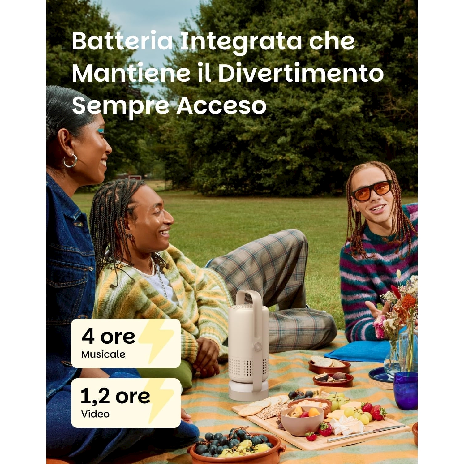 Tre persone a un picnic. Cibo, bevande e un altoparlante su una coperta. Il testo dice 'batteria integrata'.
