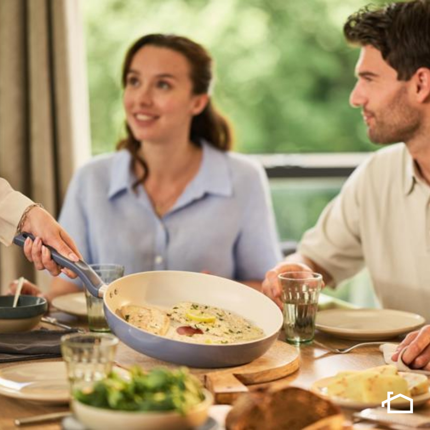 Una persona serve cibo da una padella a un tavolo da pranzo. Due persone sono sedute al tavolo.