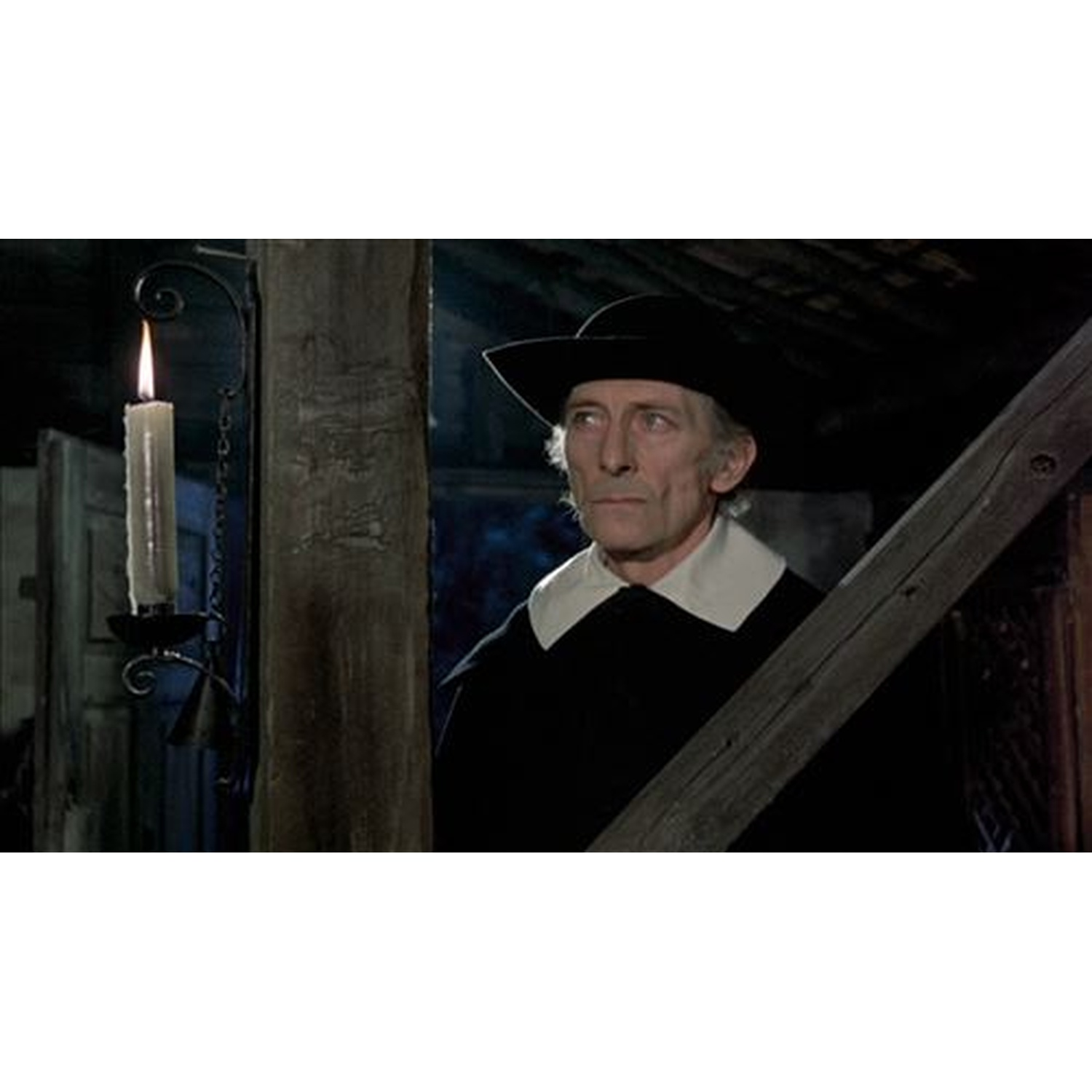 Een man in een zwarte hoed en jas, witte kraag, kijkt naar links. Een kaars brandt in een houder. Houten balken op de achtergrond.