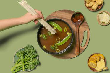Eine Hand gibt Nudeln in eine Schüssel Suppe. Brokkoli, Erbsen und andere Lebensmittel sind darum angeordnet.