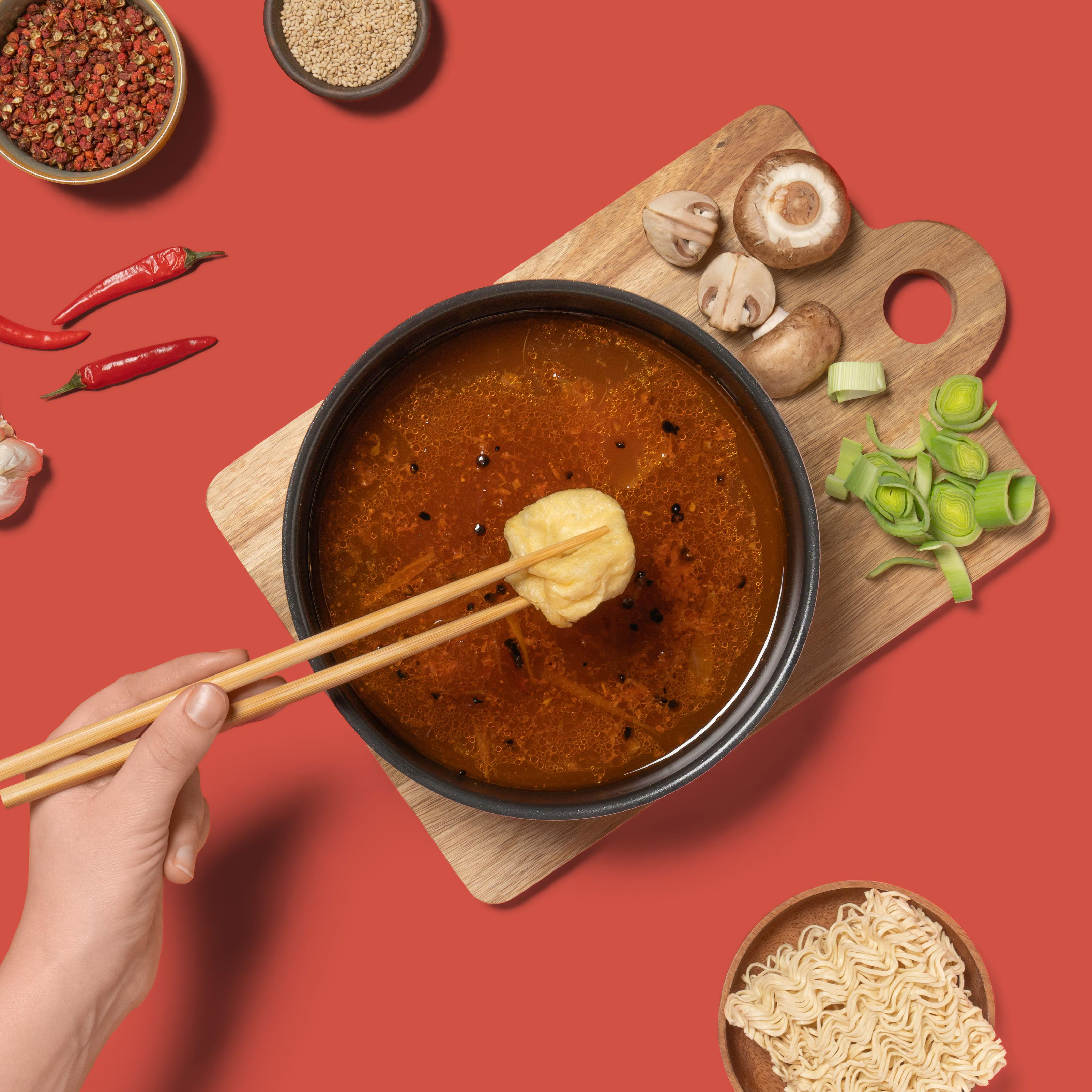 Eine Schüssel Suppe mit Stäbchen, die einen Knödel halten, umgeben von Zutaten auf rotem Grund.