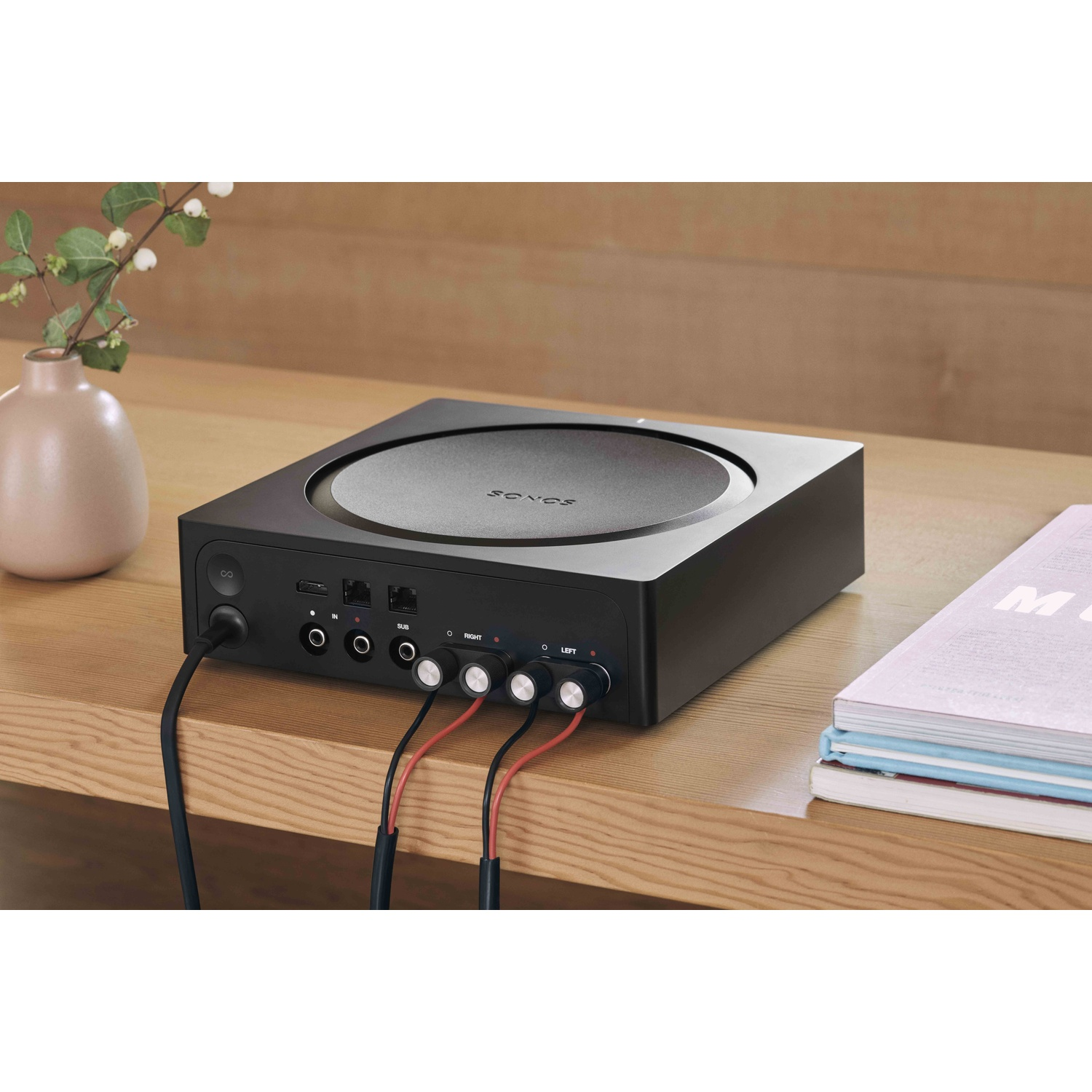 Un sistema audio Sonos nero su un tavolo di legno, con fili e un libro. Un vaso con una pianta a sinistra.