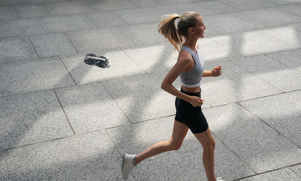 Kobieta biegnie po szarej, kafelkowanej powierzchni. Czarny dron unosi się w pobliżu. Ma na sobie szary top, czarne szorty i białe buty.