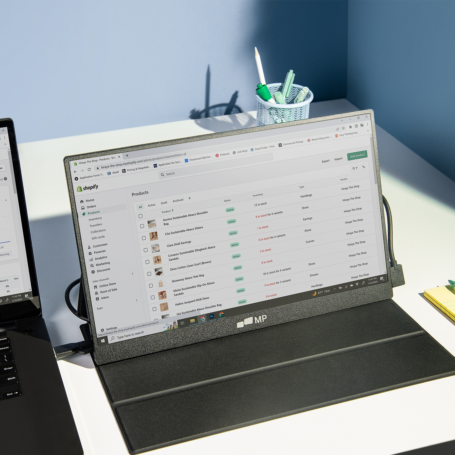 A laptop displays a Shopify products page. A secondary monitor shows the same. A pen holder and a laptop are on the table.