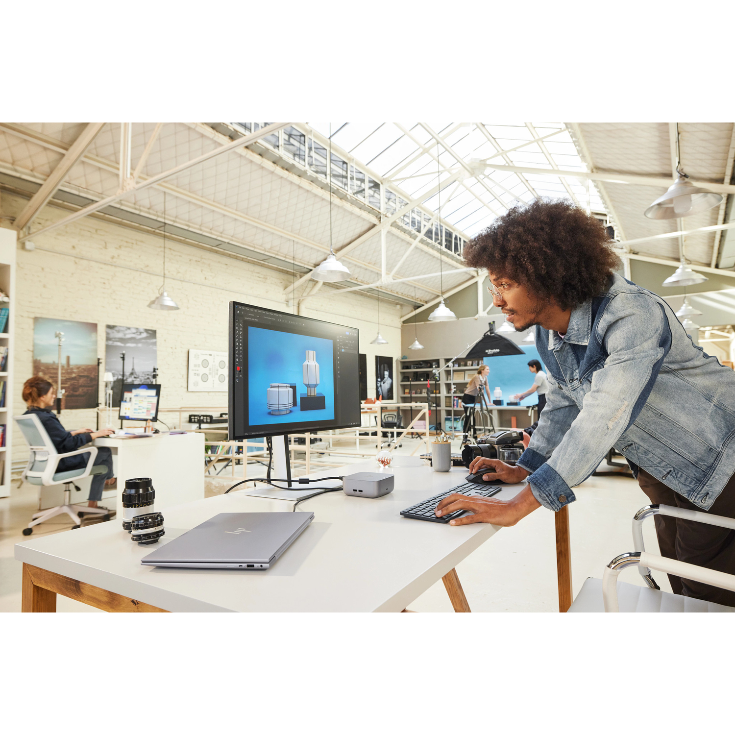 A man with afro works on a computer. A laptop, lenses, and a monitor are on the table. Other people work in the background.