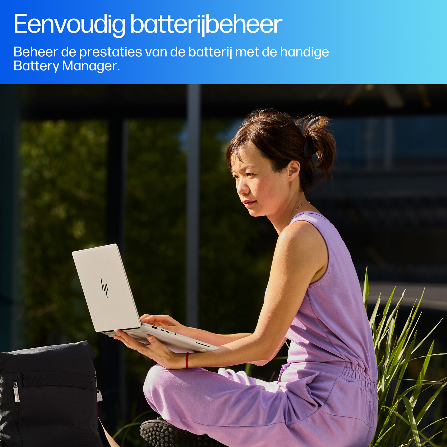 A woman in a purple jumpsuit sits outdoors with a laptop. A black bag is beside her. Green plants and a building are in the background.