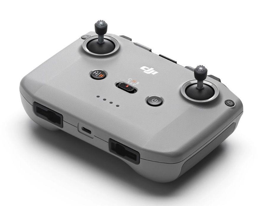 Szary kontroler drona z dwoma joystickami, przyciskami i portami. Posiada logo DJI i przycisk zasilania. Białe tło.