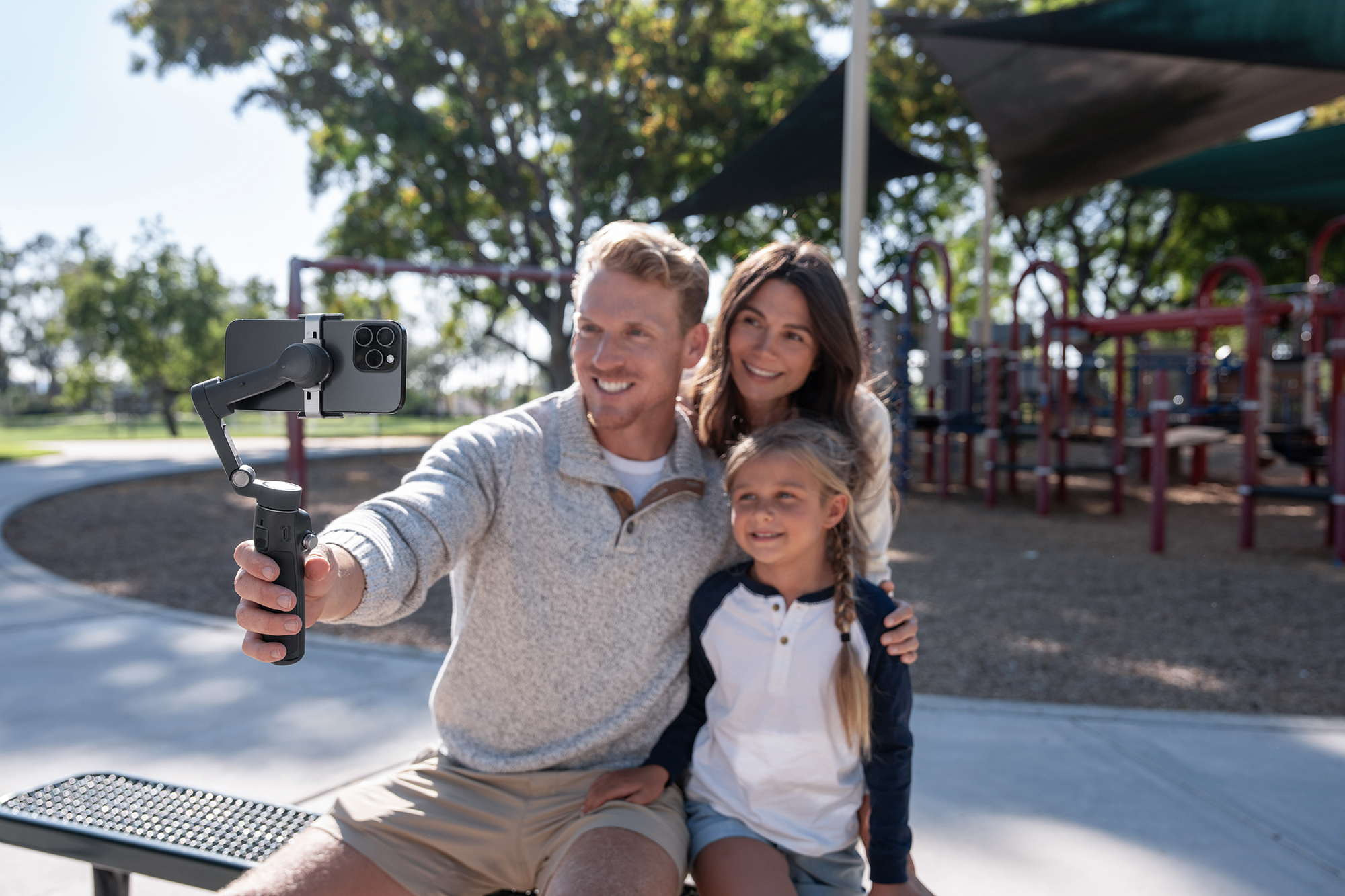 Üç kişilik bir aile, parkta bir gimbal üzerinde telefonla selfie çekiyor. Oyun alanı ekipmanlarının önünde gülümsüyorlar.