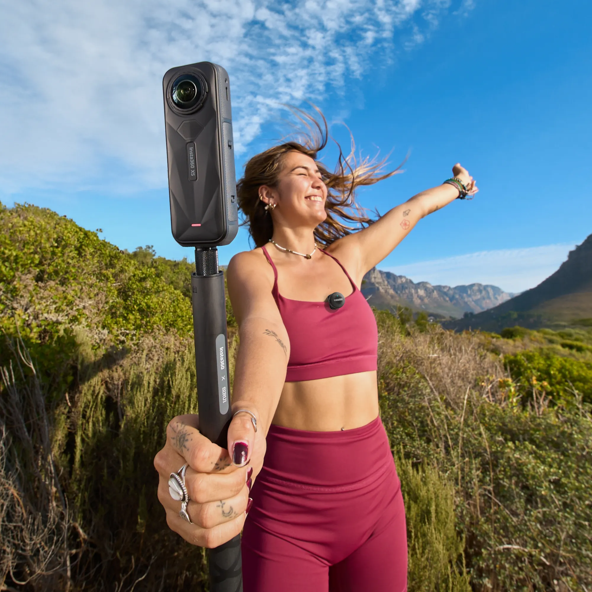 Frau hält 360-Kamera, lächelt, vor blauem Himmel. Berge und grünes Laub im Hintergrund.