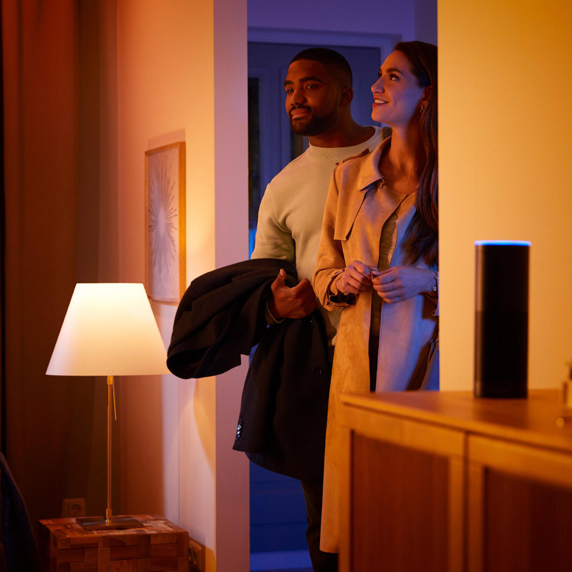 Ein Paar steht in einer Tür. Der Mann hält einen Mantel. Eine Lampe leuchtet. Ein intelligenter Lautsprecher steht auf einem Holzschrank.