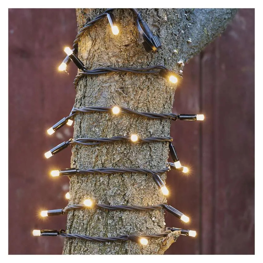 Egy fatörzs, amelyet apró, meleg fehér fényekkel díszített girland borít. A háttér elmosódott barna színű.
