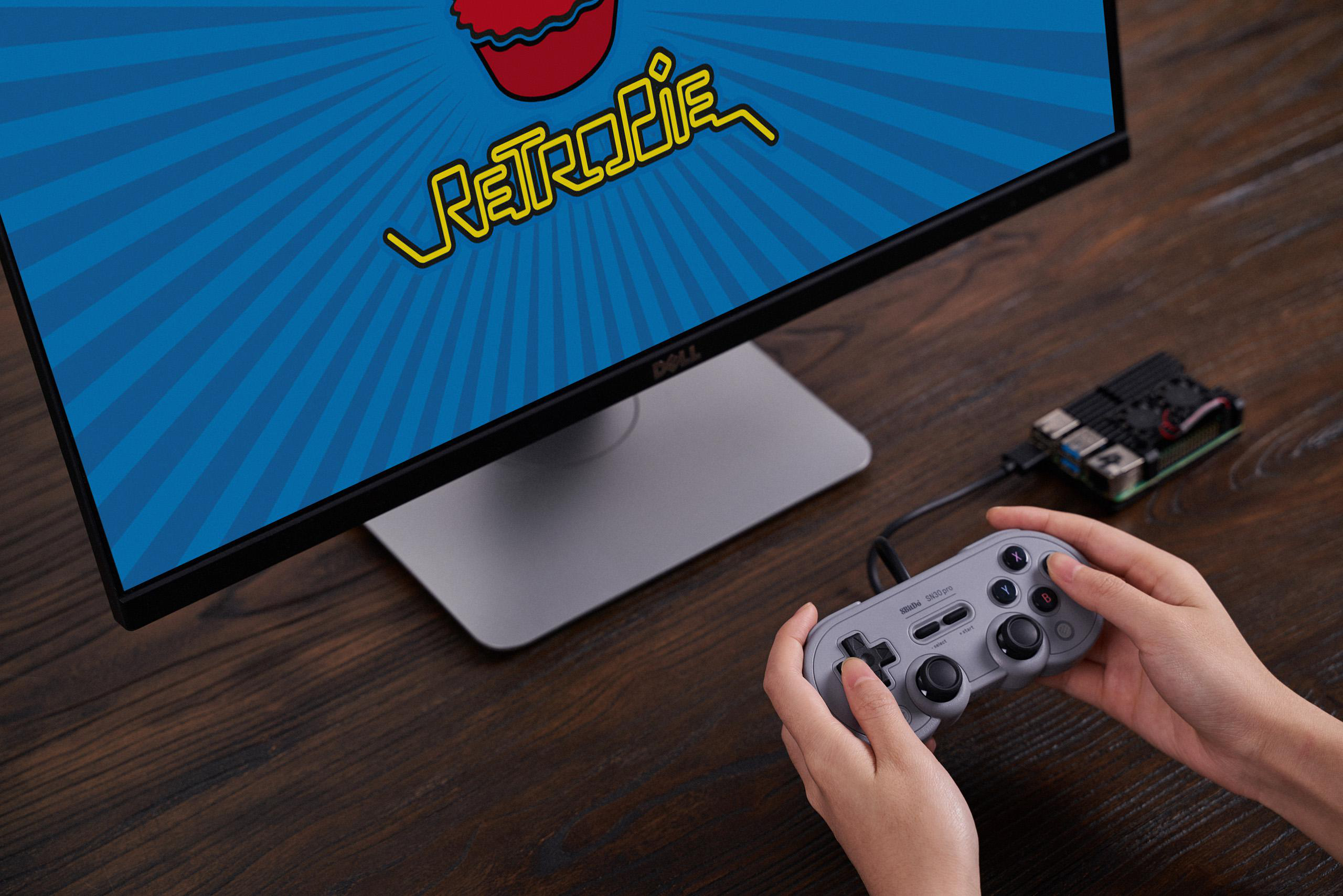 Hände halten einen grauen Gamecontroller, der mit einem Bildschirm verbunden ist, der ein blaues RetroPie-Logo zeigt.
