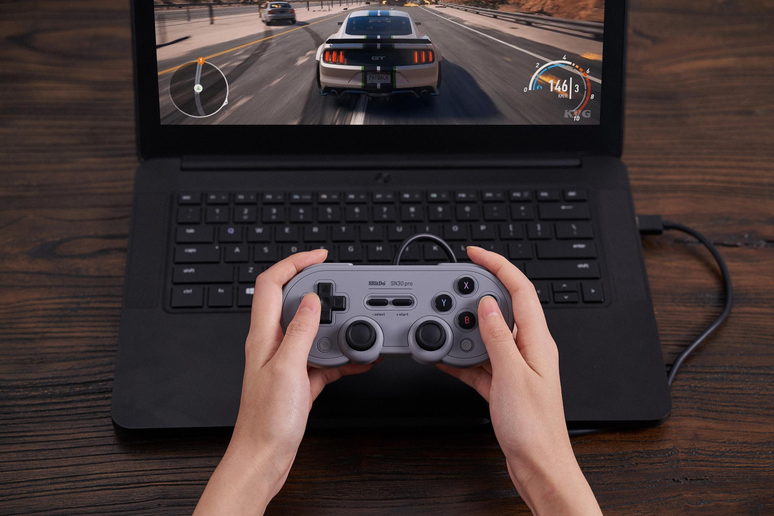 Hände halten einen grauen Controller und spielen ein Rennspiel auf einem Laptop. Das Spiel zeigt ein weißes Auto.