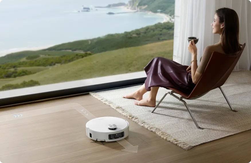 Frau mit Kaffee, Roboter-Staubsauger reinigt. Malerische Aussicht auf Meer und Hügel durch ein großes Fenster. Entspannende Szene.
