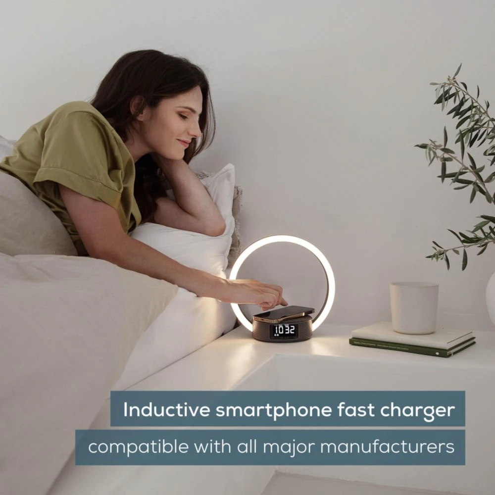 Femme au lit atteint son téléphone sur un chargeur sans fil. Une lumière annulaire est derrière le téléphone. L'horloge affiche 10:32.