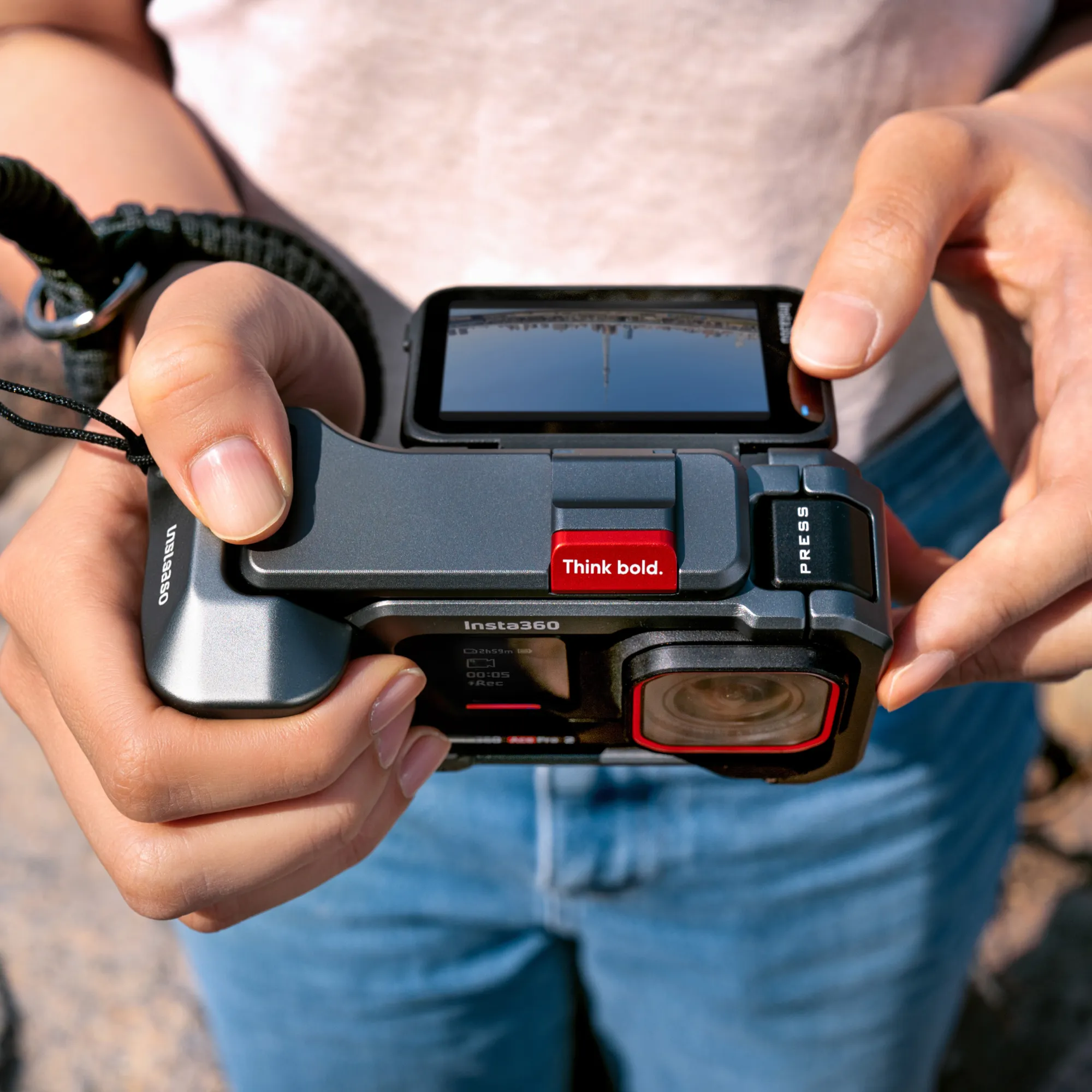 Hände halten eine graue Insta360-Kamera. Die Kamera hat einen roten Knopf mit der Aufschrift 'Think bold'. Jeans sind sichtbar.