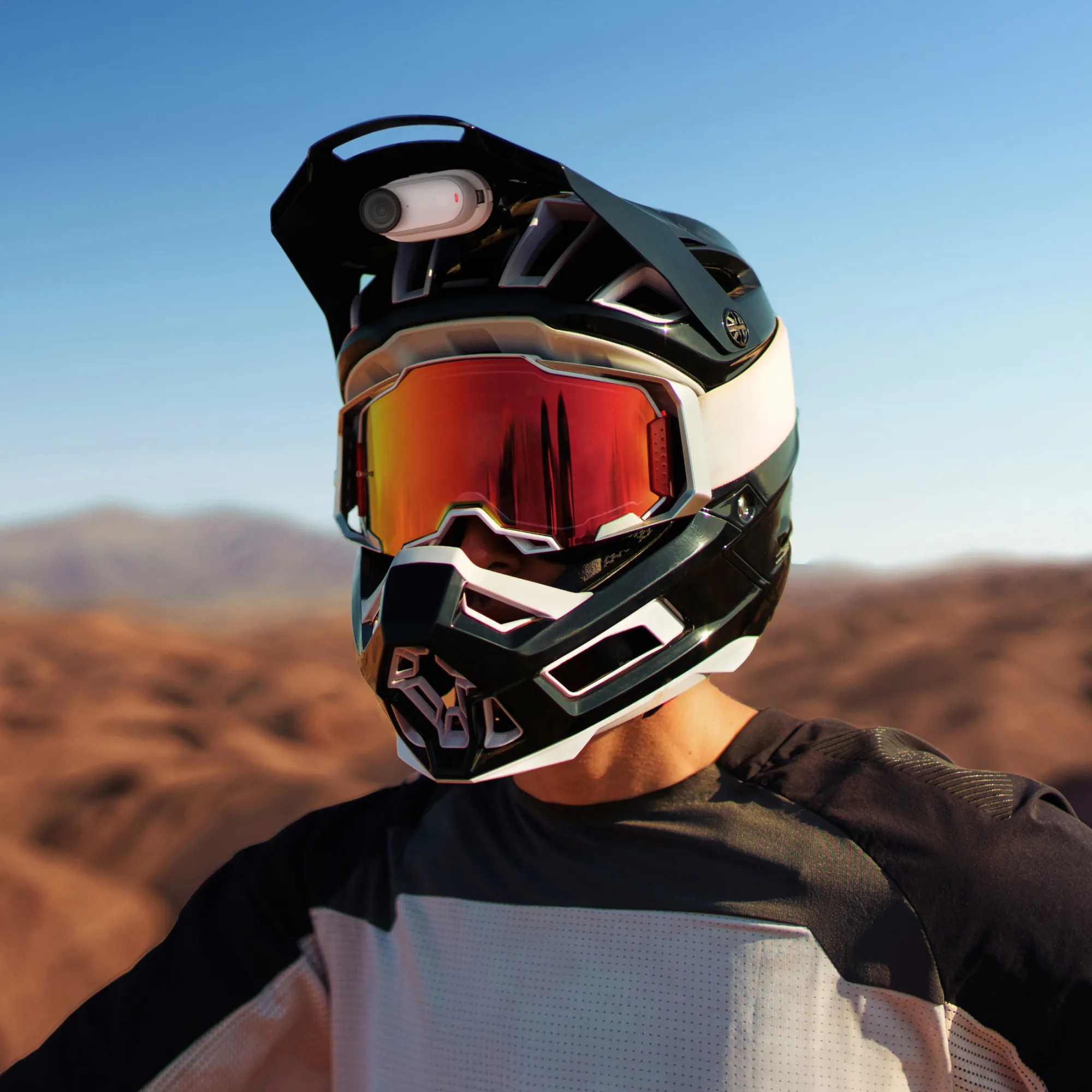Eine Person trägt einen schwarz-weißen Helm mit rot getönten Brillen. Eine Kamera ist am Helm befestigt. Der Hintergrund ist eine Wüste.