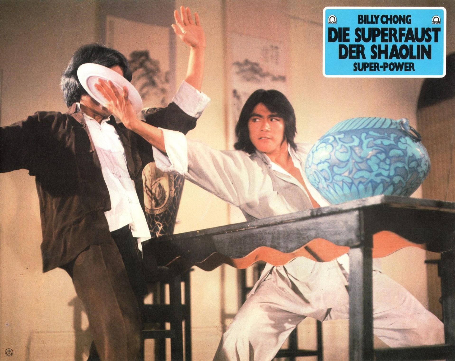 Ein Mann wird mit einem Teller getroffen. Ein anderer Mann hebt die Hand. Eine blaue Vase steht auf einem Tisch. Filmtitel oben.